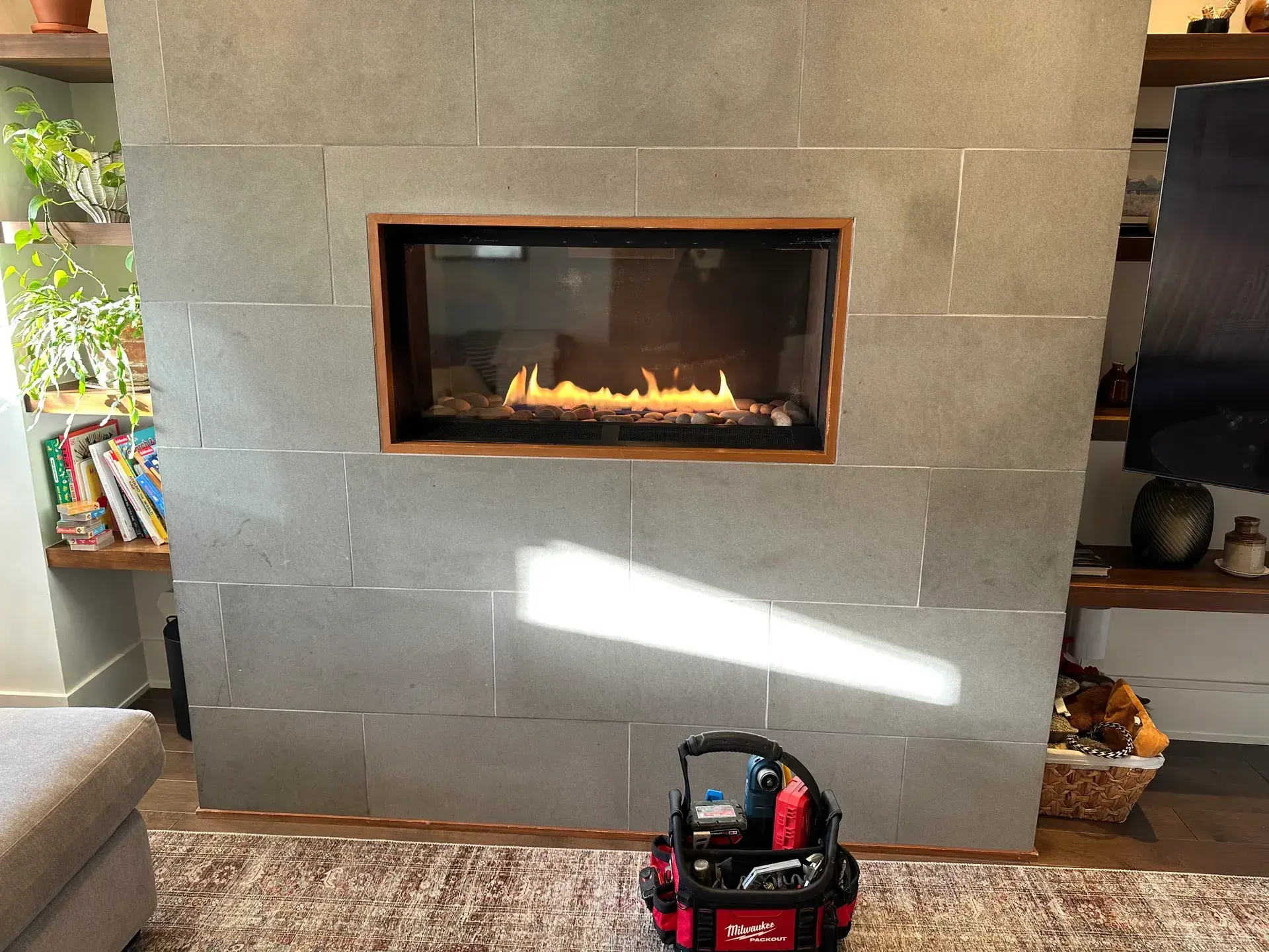 Modern linear gas fireplace burning in a floor-to-ceiling grey tile feature wall, tool bag on the rug from a live service call