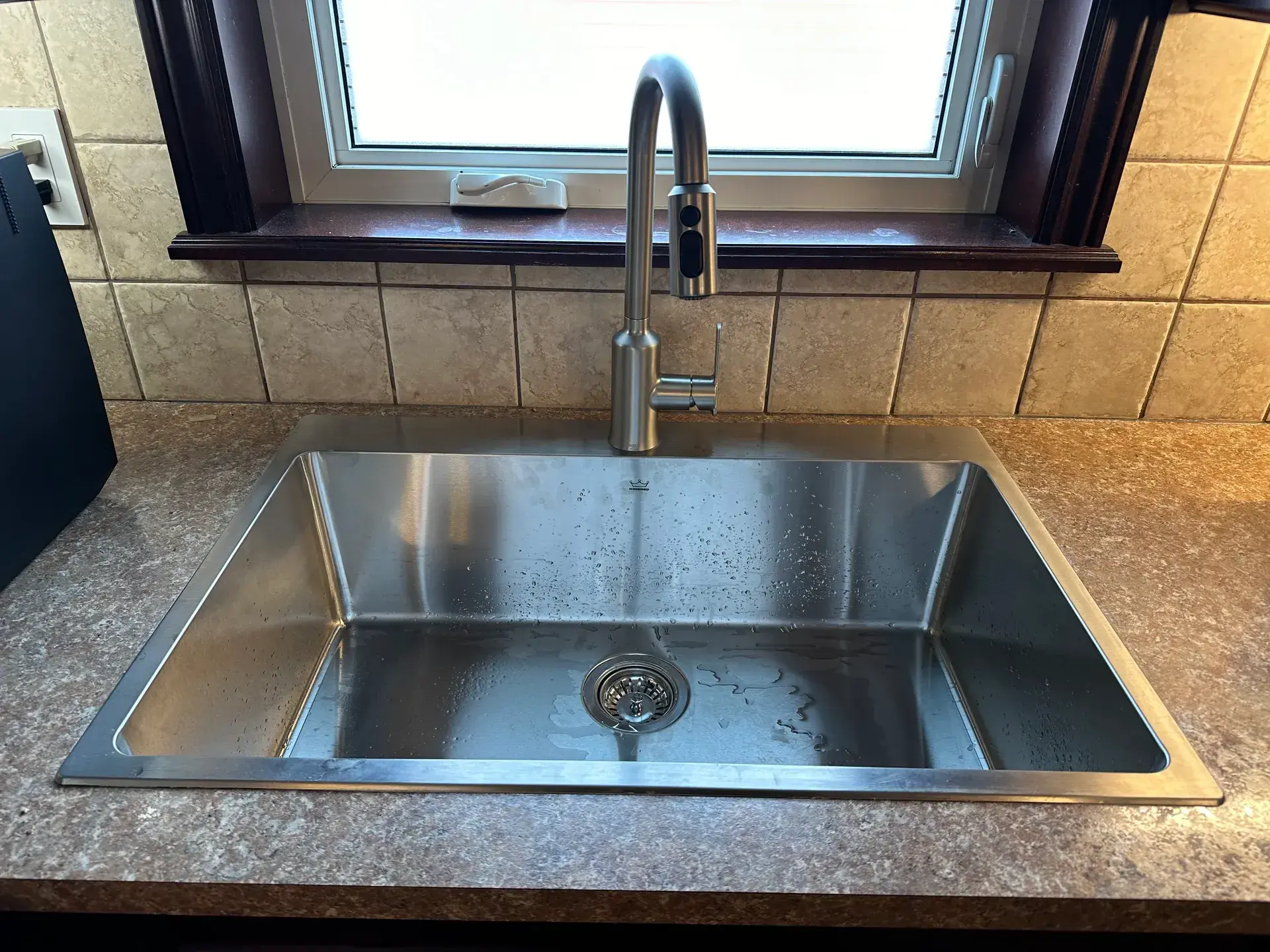Newly installed stainless kitchen sink and brushed-nickel pull-down faucet, water droplets from the test run still in the bowl
